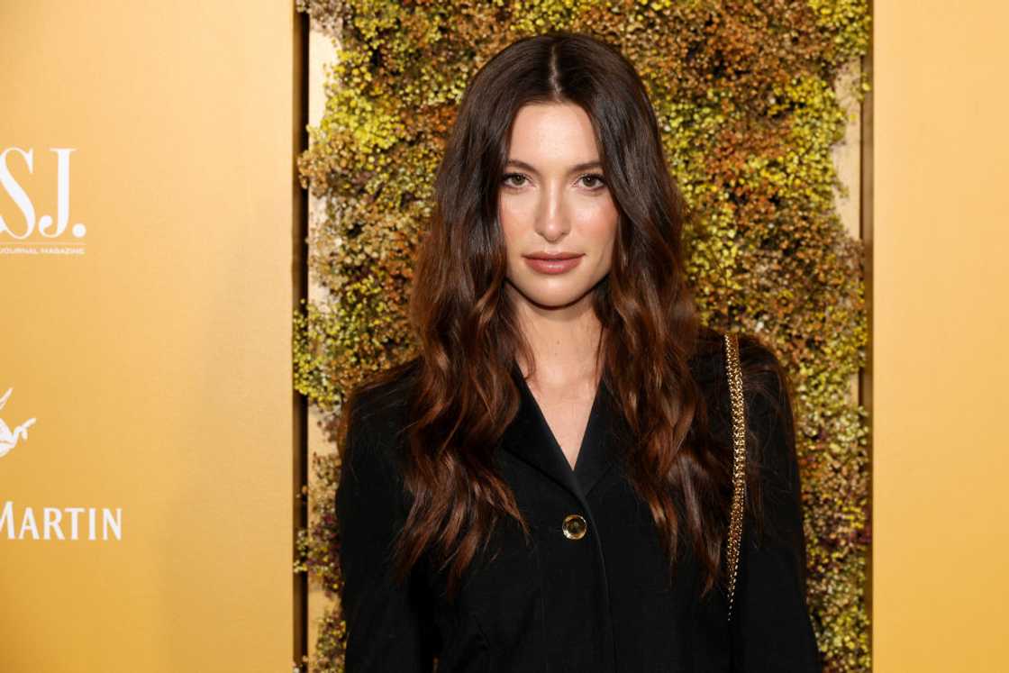 A woman with dark wavy hair poses in a black blazer against a floral backdrop.
