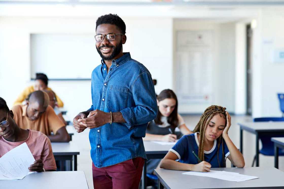 smiling young male supervisor standing amidst multi-ethnic students writing exam at desks in classroom smiling young male supervisor standing amidst multi-ethnic students writing exam at desks in classroom