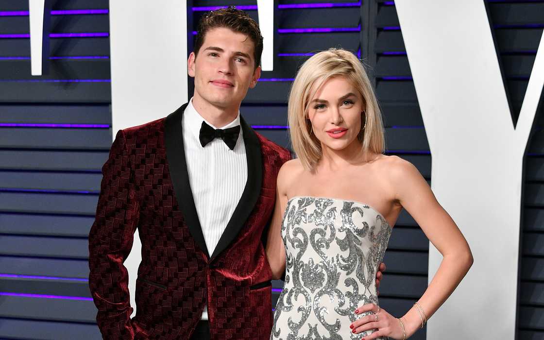 Gregg Sulkin, in a red velvet suit and Michelle Randolph, in a silver and white dress, pose on the red carpet of the 2019 Vanity Fair Oscar Party