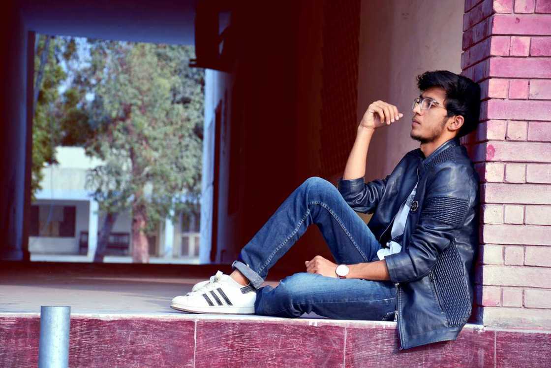 A man wearing a black leather jacket and blue jeans sitting on floor