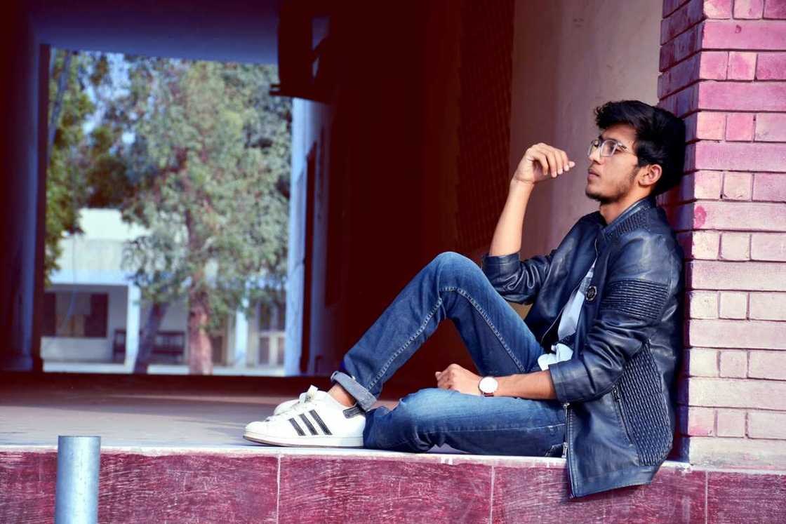 A man wearing a black leather jacket and blue jeans sitting on floor A man wearing a black leather jacket and blue jeans sitting on floor